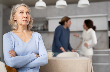 Frustrated senior woman is sitting in kitchen during quarrel between her son and his wifeの写真素材