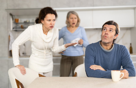 Man turned away from his wife and mother swearing at him in kitchen at homeの写真素材