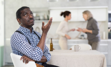 Middle-aged man sitting at the kitchen table with his back to women quarreling togetherの写真素材