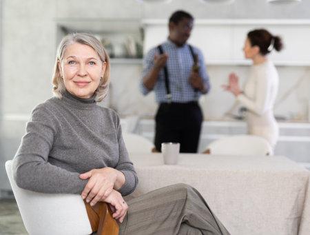 Smiling old woman sitting at the kitchen table with her back to couple quarreling togetherの写真素材