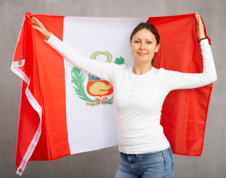 Smiling relaxed woman waving national flag of Peru while looking at camera at gray wall backgroundの写真素材