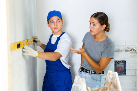 Young worker uses a spirit level on wall, consulting with a girl client
