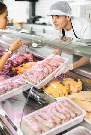 Positive young salesman demonstrating container with sausages in butcher shopの写真素材