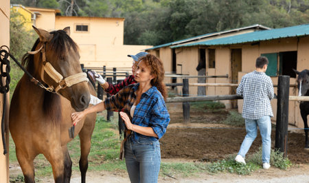 Two female workers in plaid shirt combs tethered horse in stable.