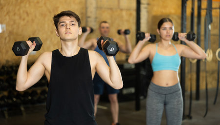 Young dedicated active strong fit man doing exercises for shoulders while pushing up dumbbells in CrossFit centerの写真素材