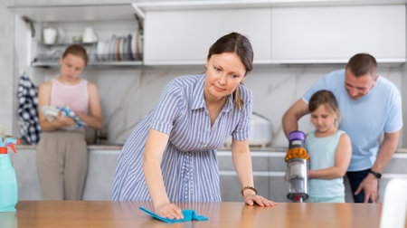 Wife cleans table with rag, husband and children clean kitchen in backgroundの写真素材