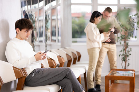 Young man browsing websites on smartphone while sitting in lobby of job center or employment office, waiting for interview or conversation with career counselorの写真素材