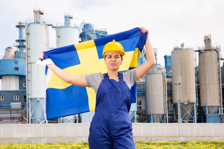 Upset disappointed young female engineer in helmet waving state flag of Sweden during strike in front of big tanks at chemical plantの写真素材