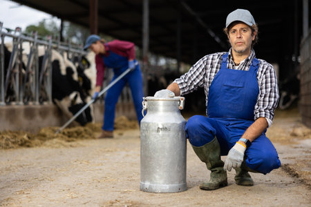 Confident hardworking man, owner of farm in overalls with metallic milk can on livestock farm in autumnの写真素材