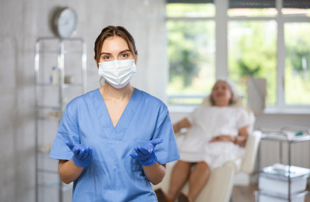 Happy doctor or nurse with mask in uniform with crossed arm, patient behind on couch.の写真素材