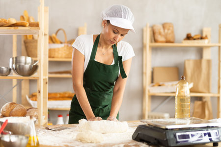 Smiling young female professional baker in apron standing at work table and kneading dough during...