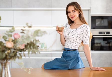 Girl is sitting on kitchen table and holding clear glass cup with coffee in handsの写真素材