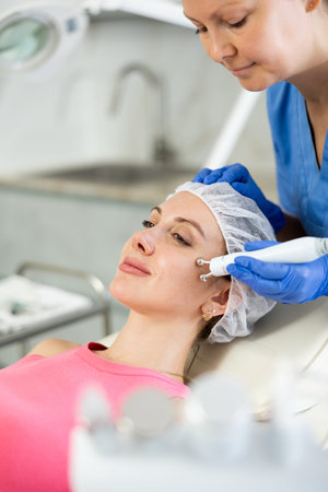 Adult woman cosmetologist doing microcurrent procedure to young woman