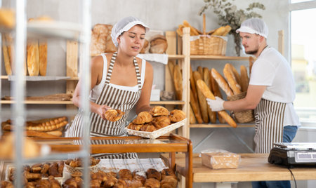 Young woman places croissants under the display while a man arranges baguettes on shelve