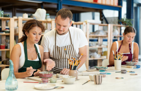 During master class, student paints clay vessel under guidance of male teacher.