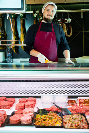 Smiling butcher behind counter with meat products in refrigerated showcaseの写真素材