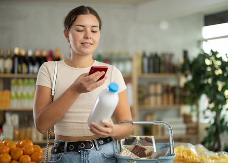 European young woman customer at shop scans QR code on milk bottle using phone cameraの写真素材