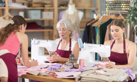Two craftsperson sew clothes, girl assistant attaches pattern to fabric.