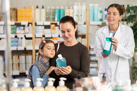 Woman with schoolgirl daughter are busy choosing mouthwash, pharmacist offer help in backgroundの写真素材