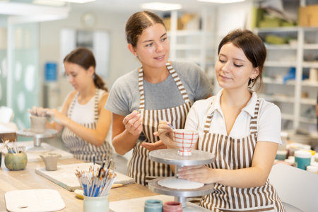 During master class, student paints clay vessel under guidance of female teacher.