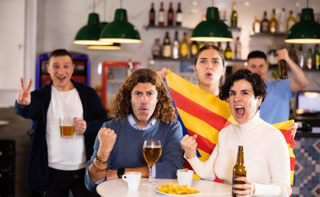 Excited sports supporters of different ages with flag of Estelada, rejoicing winning game with beer and snacks in pubの写真素材