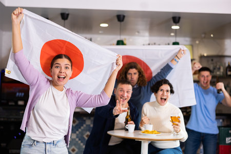Enthusiastic company of young adult sports fans waving flag of Japan and supporting favorite team with beer in pubの写真素材