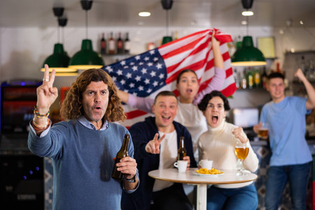 Emotional company of young adult sports fans supporting American team with state flag while resting in bar with beerの写真素材
