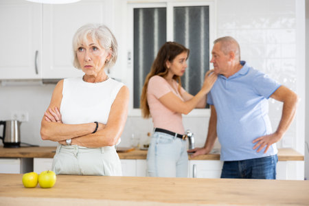 Elderly frustrated woman turned away from scolding her husband and adult daughter in kitchenの写真素材
