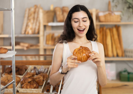 Young female customer stands with croissant in hands near window of bakery.の写真素材