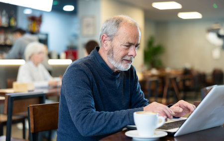 Mature man drinks coffee and works on a laptop in cafe interior