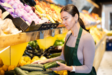 Female salesperson carefully places ripe cucumbers on counter of a grocery supermarketの写真素材