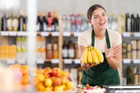 Young woman seller in store is standing near fruit counter, selling bananas.の写真素材