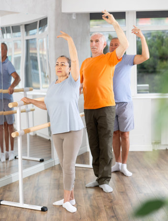 Ordinary elderly people stand in third position near ballet barre during group training in dance...