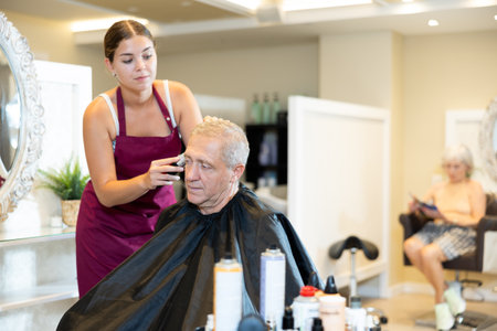 Young woman hairdresser performs haircut for elderly man using hair clipperの写真素材