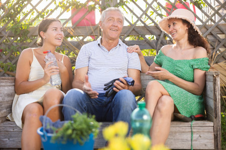 Woman in straw hat is sitting on wooden bench and chatting sweetly with sister and senior fatherの写真素材