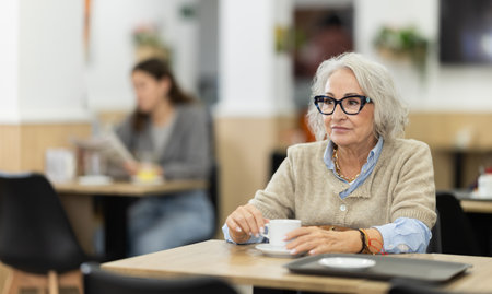Aged european woman drinking fresh coffee at table in cafeの写真素材