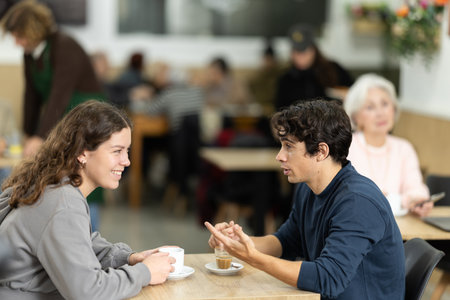 Young couple drinking coffee and chatting while sitting at table in cozy cafeの写真素材