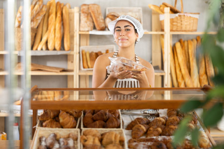 Young saleswoman holds out croissants in plastic bag in bakery