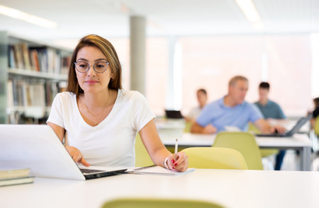 Portrait of adult latino woman studying at library using books and laptop