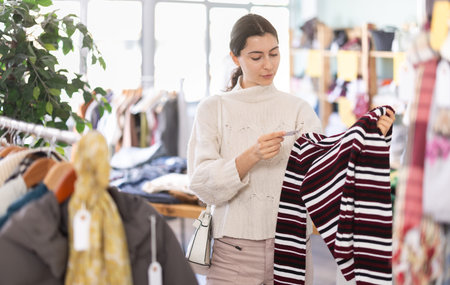 Fashionable young girl standing near shelves with clothes in modern store and choosing warm sweater