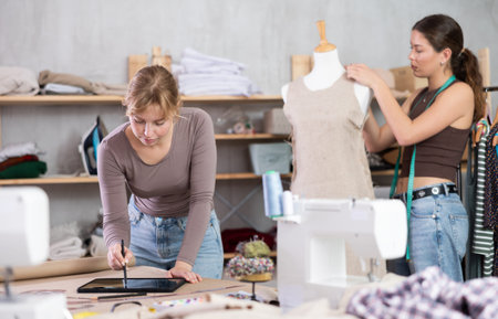 Dressmaker sketching dress on tablet while colleague working with mannequin