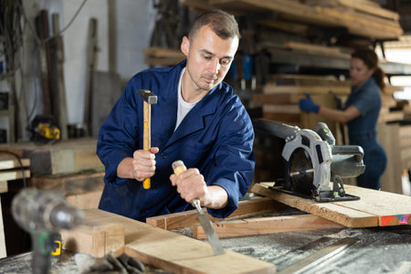 Male employee of carpentry workshop performs preparatory work on board with hammer and chisel