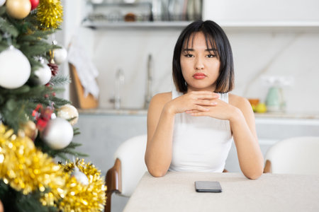 Sad young Asian girl waiting for phone call sitting at table by Xmas treeの写真素材