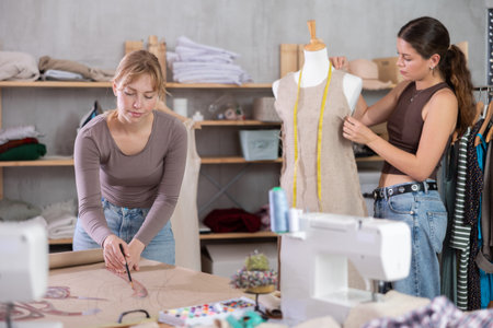 Female dressmaker drawing paper pattern as colleague adjusting sample on mannequin