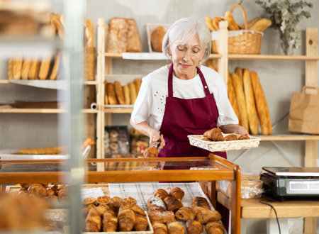 Mature woman employee puts croissants in window, arranges display of goods at bakery.の写真素材