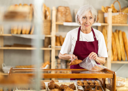 Mature saleswoman putting croissants in plastic bag in bakeryの写真素材