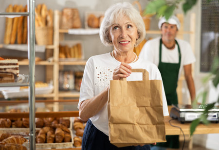Senior woman bakery customer stands in sales area with paper bag in handsの写真素材