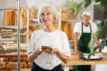 Senior female client takes piece of puff pastry cake from shelf of showcase, examines productsの写真素材