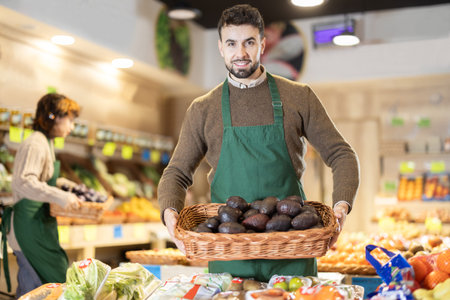 Young male seller holding basket of avocados standing by counter in grocery marketの写真素材