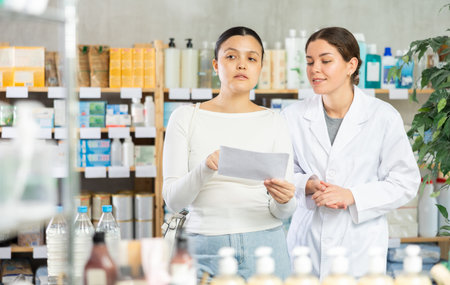 Portrait of young woman pharmacist helping asian female client with paper in hand looking for something or choosingの写真素材